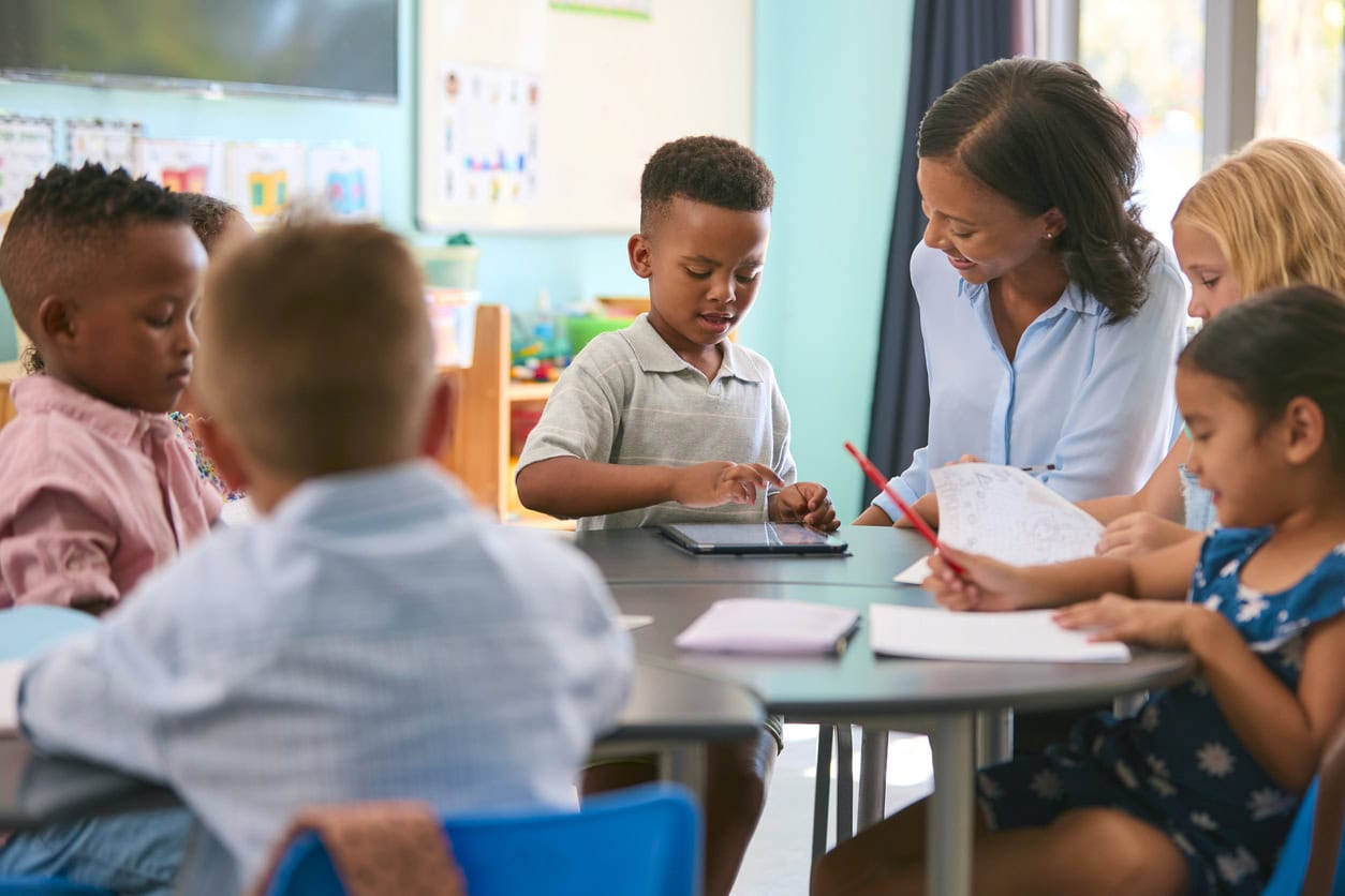 Female Primary Or Elementary School Teacher Helping Students With Digital Tablet At Table In Class