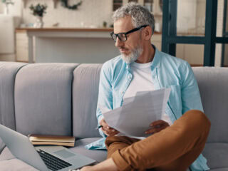 Busy senior man checking the papers and using laptop while sitting on the sofa at home